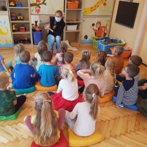 pokaż obrazek - Sala przedszkolna. Starszaki siedzą na kolorowych poduszkach zwrócone plecami do widza. Naprzeciwko nich pani siedzi na małym krzesełku i pokazuje ilustrację w książce. Dzieci przyglądają się z zainteresowaniem.