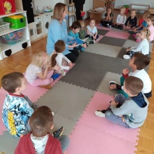 pokaż obrazek - W sali przedszkolnej na różowych i szarych matach w kształcie prostokątów siedzi grupa skupionych dzieci. Pomiędzy nimi kobieta w niebieskiej bluzce trzymająca książkę. Z tyłu zabawki i kolorowe meble.