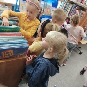 pokaż obrazek - Cztery dziewczynki i chłopiec stoją pomiędzy regałami. Dziewczynka w żółtej koszulce i różowej opasce wertuje książki. Jej dwie koleżanki przyglądają się z zainteresowaniem okładkom.