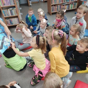 pokaż obrazek -  Grupa kilkuletnich chłopców i dziewczynek pochyla się nad pokazywana przez panią książką. Pani  wskazuje na ilustrację palcem a jedna z dziewczynek przytrzymuje brzeg oglądanej książki. W tle regały z książkami.