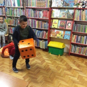 pokaż obrazek - Sala biblioteczna. Na pierwszym planie chłopiec z ogromną pomarańczową kostką wykonuje rzut. W tle regały z książkami oraz dwie dziewczynki.