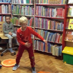 pokaż obrazek - Sala biblioteczna. Na pierwszym planie chłopiec w czerwonej koszulce patrzy za rzuconą wcześniej kostką. W tle regały z książkami oraz dwie dziewczynki.