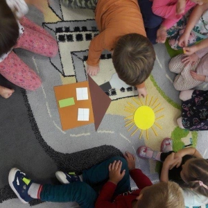 pokaż obrazek - Dzieci siedzą na dywanie. Chłopiec kończy układać obrazek na dywanie. Obrazek przedstawia domek i słoneczko.