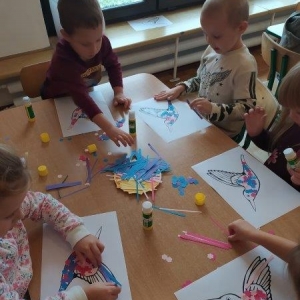 pokaż obrazek - W sali przedszkolnej przy stoliku siedzą kilkuletnie dzieci, które wyklejają ptaszki kolorowymi kwiatkami i paskami. 