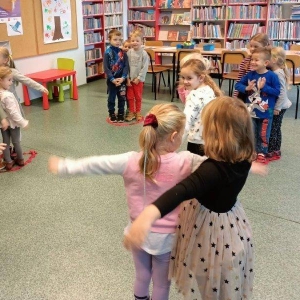 pokaż obrazek - W sali bibliotecznej w czerwonych kręgach stoją ustawione w trójki kilkuletnie uśmiechnięte dzieci. Niektóre dzieci wyciągają ręce na bok. Z tyłu regały biblioteczne.