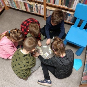 pokaż obrazek - Dwie dziewczynki i trzech chłopców w wieku wczesnoszkolnym siedzi na podłodze w bibliotece. Przed nimi puzzle z zającem. Dzieci próbują je ułożyć. W prawym górnym rogu niebieskie dziecięce krzesełko. Z tyłu widać fragmenty regałów z książkami.