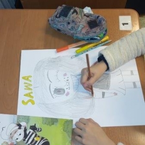 pokaż obrazek - Na biurku kartka z rysunkiem i żółtym napisem, piórnik i komiks. Widać ręce rysującej dziewczynki, która w prawej dłoni trzyma ołówek . W górnej części zdjęcia kawałek drugiego biurka z brzegiem kartki i komiksu.  