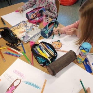 pokaż obrazek - Przy stoliku dwie dziewczynki rysują coś mazakami. Obok nich porozkładane piórniki, mazaki, strugaczki, ołówek i gumka. W prawym dolnym rogu dziecięca ręka z zieloną kredką.