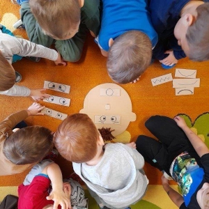 pokaż obrazek - Grupa przedszkolaków pochyla się nad konturem głowy postaci. Dzieci starają się  dopasować minę do emocji. 