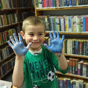 pokaż obrazek - Uśmiechnięty chłopiec w zielonej koszulce pokazuje wnętrze swoich dłoni, pomalowanych na niebiesko. W tle regały z książkami.