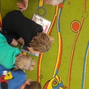 pokaż obrazek - Dzieci siedzą na dywanie pochylone nad książką. Chłopiec trzyma w ręce specjalną latarkę, którą porusza w książce.