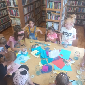 pokaż obrazek - Sala biblioteczna. Grupa dziewczynek dziedzi wokół dużego stołu, na którym znajdują się materiały plastyczne: słoiki, piasek, muszelki, niebieska bibułka, klej, kartki papieru. W tle regały z książkami.