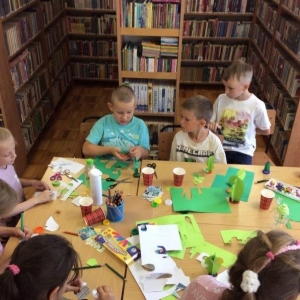 pokaż obrazek - Biblioteka.Grupa dzieci siedzi przy stolikach i wykonuje pracę plastyczną - papierowego kaktusa. Na stole kolorowe kartki papieru, nożyczki, klej, pisaki. W tle regały z książkami.