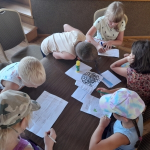 pokaż obrazek - Szóstka dzieci /cztery dziewczynki i dwóch chłopców/siedzi przy stoliku i rozwiązują zagadki. Dziewczynki rozwiązują zagadki a chłopcy nachylają się nad kartkami. Na stoliku klej, kartki z zadaniami i dwie koperty.