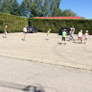 pokaż obrazek - Parking za biblioteką. Grupa dzieci gra w papierową piłkę nożną. W tle dwa samochody, żywopłot oraz drzewa.