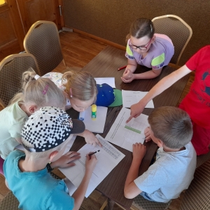 pokaż obrazek - Szóstka dzieci /trzy dziewczynki i trzech chłopców/siedzi przy stoliku i rozwiązują zagadki. Chłopiec w czerwonej koszulce wskazuje palcem na kartkę z zadaniami. Na stoliku klej, trzy kartki z zadaniami oraz czerwona kredka.