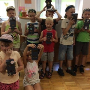 pokaż obrazek - Sala biblioteczna. Ośmioosobowa grupa dzieci  trzyma w rękach wykonane przez siebie biblio-boty. W tle okna.