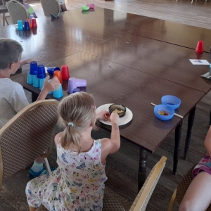 pokaż obrazek - Wokół dużego stołu siedzą dzieci podzielone na dwie drużyny. Wykonują zadanie na czas. Na stole znajdują się: kolorowe plastikowe kubki, miseczki, pałeczki, pojemniki z różnymi ziarnami do posegregowania. Dwóch chłopców układa kubki, dwie dziewczynki rozdzielają ziarna. Pozostałe dzieci czekają na swoją kolej.