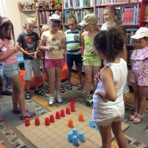 pokaż obrazek - Pomieszczenie biblioteczne. Grupa dzieci – chłopców i dziewczynek stoi wokół planszy do kodowania, na której ułożone są kolorowe kubeczki. Dzieci starają się odgadnąć zakodowany kształt. W tle regały z książkami.