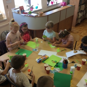 pokaż obrazek - Biblioteka.Grupa dzieci siedzi przy stolikach i wykonuje pracę plastyczną - papierowego kaktusa. Na stole kolorowe kartki papieru, nożyczki, klej, pisaki, papierowe kubeczki. W tle lada biblioteczna i okna.