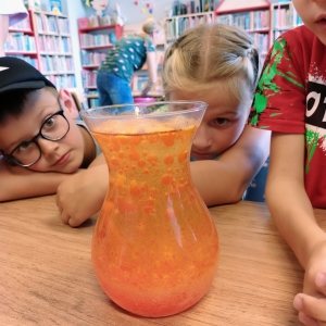 pokaż obrazek - Biblioteka. Na stoliku stoi przeźroczysty wazon, a w nim buzująca pomarańczowa ciecz. Przyglądają się temu dwaj chłopcy i dziewczynka. W tle regały z książkami.