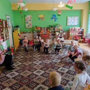 pokaż obrazek - Zdjęcie przedstawia salę przedszkolną, w której na krzesełkach siedzą dzieci, twarzami skierowane w stronę bibliotekarki trzymającej książkę. W tle regały z zabawkami i stoliki.