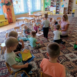 pokaż obrazek - Zdjęcie przedstawia salę przedszkolną, w której na dywanie siedzą dzieci, oglądają położone przed sobą lub trzymane w rękach książeczki. W tle widać kolorowe regały z zabawkami i stoliki.