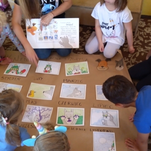 pokaż obrazek - Zdjęcie przedstawia grupę kilkuletnich dzieci siedzących w kółku na dywanie. Przed nimi rozłożony szary papier z wypisanymi nazwami miesięcy, a na nim kolorowe ilustracji. Pani trzyma książkę na kolanach i pokazuje dzieciom ilustracje. Wspólnie sprawdzają właściwą kolejność.