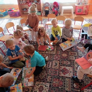 pokaż obrazek - Zdjęcie przedstawia salę przedszkolną, w której dzieci siedząc na dywanie i krzesełkach oglądają książki. Za nimi w sali znajdują się zabawki: lalki, wózki, klocki i plastikowa kuchenka.