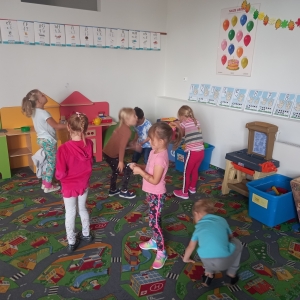 pokaż obrazek - Na dywanie stoją dzieci. Każde dziecko trzyma piórko, w które dmucha. W tle podłoga i szafki.